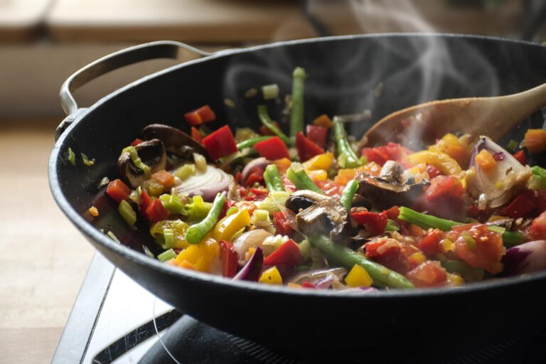 A black frying pan on a stovetop contains a colorful mix of sautéed vegetables, including green beans, red and yellow bell peppers, mushrooms, onions, and diced tomatoes. Steam is rising from the pan, indicating the food is hot and freshly cooked. A wooden spoon is stirring the vegetables, and the background shows a wooden countertop with a warm, natural light setting