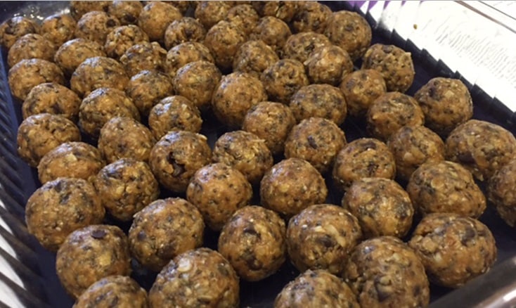 Tray filled with rows of uniform, round energy bites or protein balls made from a mixture of oats, seeds, and other ingredients, giving them a textured appearance. The balls are arranged neatly in a grid pattern inside a rectangular container.