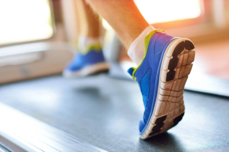 Close-up of a person running on a treadmill, focusing on the lower legs and feet. The individual is wearing bright blue athletic shoes with white soles and green accents, along with white socks. The treadmill belt is visible beneath the shoes, and the background is softly lit, suggesting an indoor fitness setting