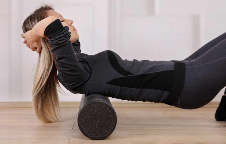 Person lying on a wooden floor using a black foam roller under the upper back, wearing a dark gray and black athletic outfit with long sleeves and leggings.