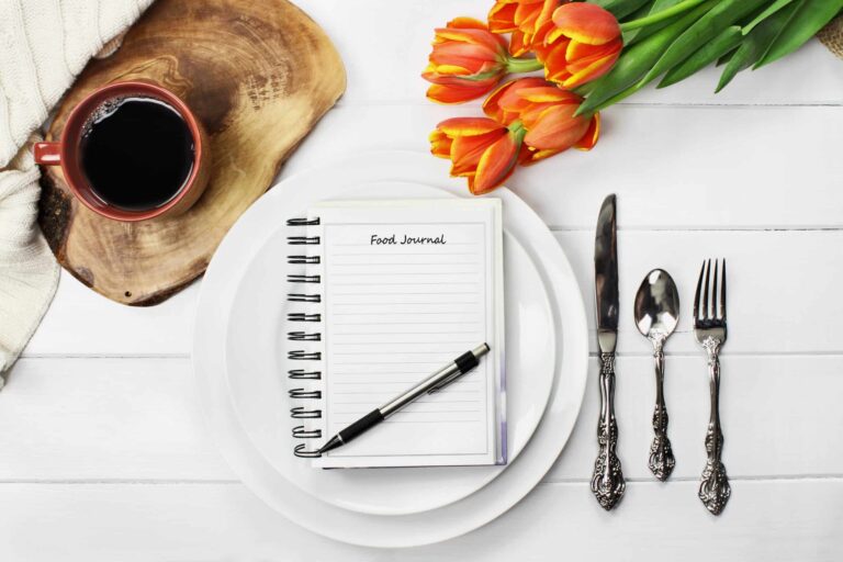A white table setting with a spiral notebook labeled “Food Journal” placed on a stack of white plates, a pen resting on the notebook, a cup of black coffee on a wooden board to the left, a bouquet of orange tulips at the top right, and a knife, spoon, and fork neatly arranged on the right side.