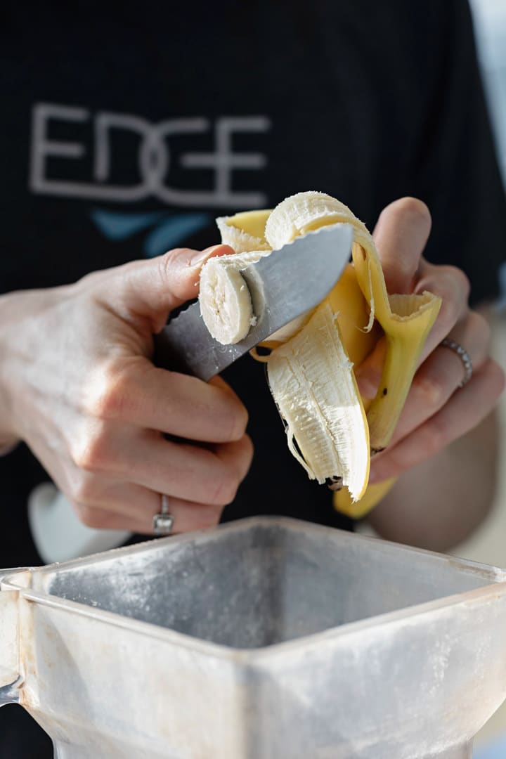 Close-up of hands slicing a banana with a knife over a blender container, with part of a shirt showing the word “EDGE” in the background.