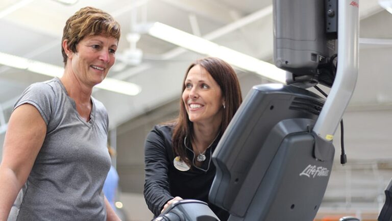 Two people standing near an elliptical machine in a gym, with one person appearing to explain or demonstrate the equipment to the other.