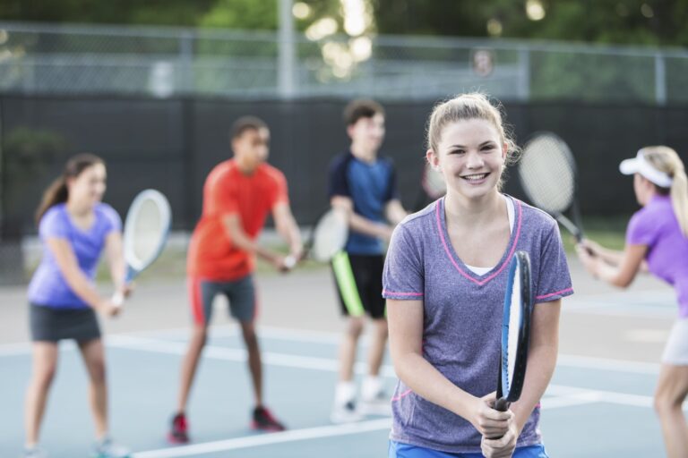 Multi-ethnic group of teenagers (14 to 16 years) at tennis clinic with instructor (30s). Focus on girl in foreground (15 years).