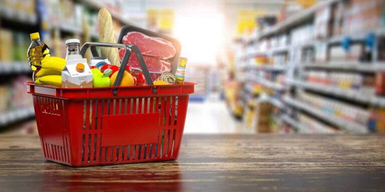 A red shopping basket filled with various grocery items is placed on a wooden surface in a supermarket aisle. The basket contains bananas, a loaf of bread, a carton of orange juice, a bottle of cooking oil, packaged meat, canned goods, and other assorted food items. The background shows shelves stocked with products, slightly blurred to emphasize the basket in the foreground.