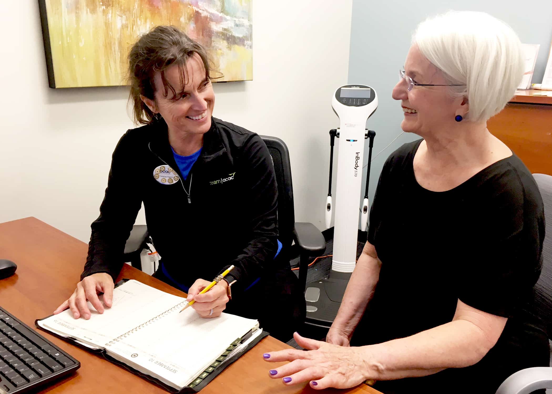 Two people sitting at a desk in an office setting, engaged in a consultation. One person is writing in an open notebook with a pencil, while the other is seated nearby with hands resting on the desk. A computer monitor is partially visible on the left side, and a body composition scale is positioned in the background. The room has light-colored walls, a framed abstract painting, and standard office furniture.