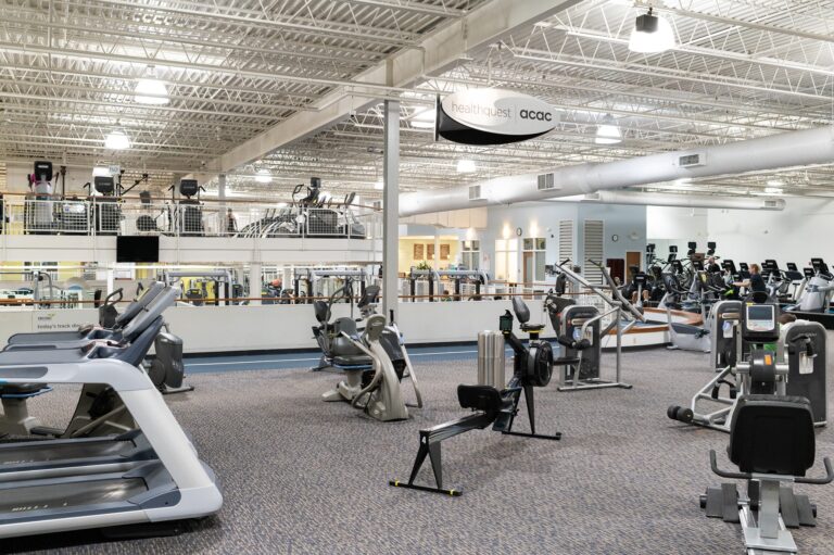 Spacious fitness center interior with various exercise equipment including treadmills, stationary bikes, rowing machines, and weight machines. The gym has high ceilings with exposed beams, bright lighting, and a mezzanine level featuring additional cardio machines. A sign overhead reads “healthquest acac.”
