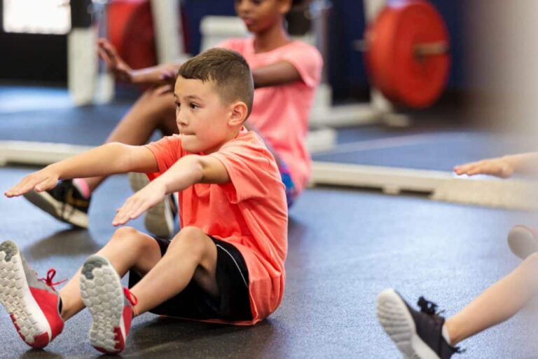 Child performing a seated exercise with arms extended forward in a gym setting, wearing athletic clothes and sneakers. Other children are exercising in the background near weightlifting equipment.