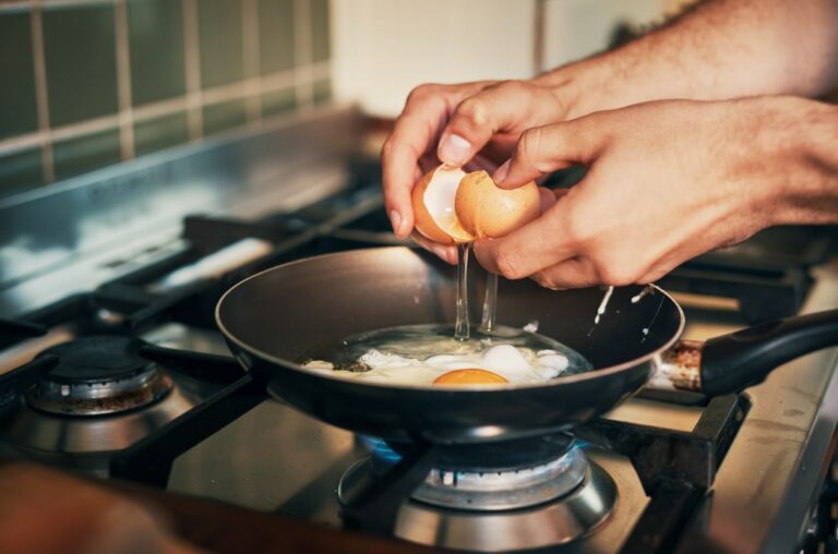 Close-up of hands cracking an egg into a frying pan on a gas stove, with another egg already cooking. The kitchen setting includes metal burners and a tiled backsplash in the background.
