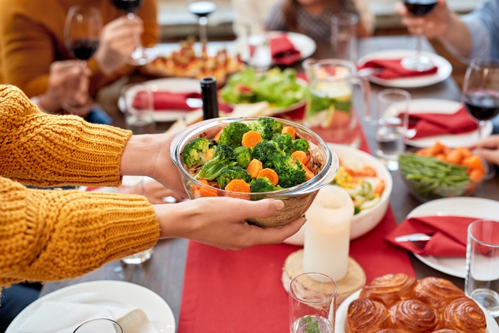 Person holding a glass bowl filled with steamed broccoli and sliced carrots over a dining table set for a meal, with plates, glasses, candles, and various dishes including salads and pastries arranged on a red table runner.