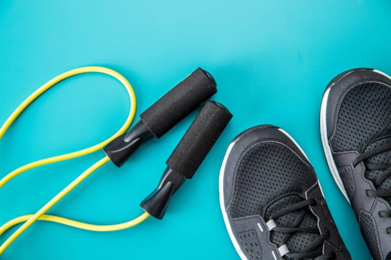 A pair of black athletic shoes placed next to a yellow jump rope with black foam handles on a bright turquoise background.