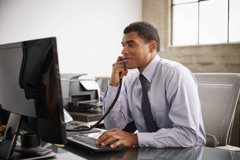 Person wearing a light blue dress shirt and a dark polka-dot tie is seated at a desk in an office setting, using a desktop computer while holding a corded phone to the ear. The desk has a keyboard, mouse, and other office items, and a printer is visible in the background near a window.