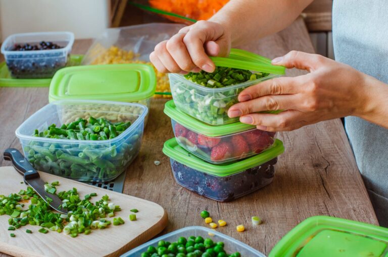 Hands are placing lids on plastic food containers filled with various fresh ingredients, including chopped green onions, strawberries, and peas. The containers are arranged on a wooden countertop alongside a cutting board with a knife and more chopped green onions. Other containers in the background hold beans and corn, suggesting meal prep or food storage