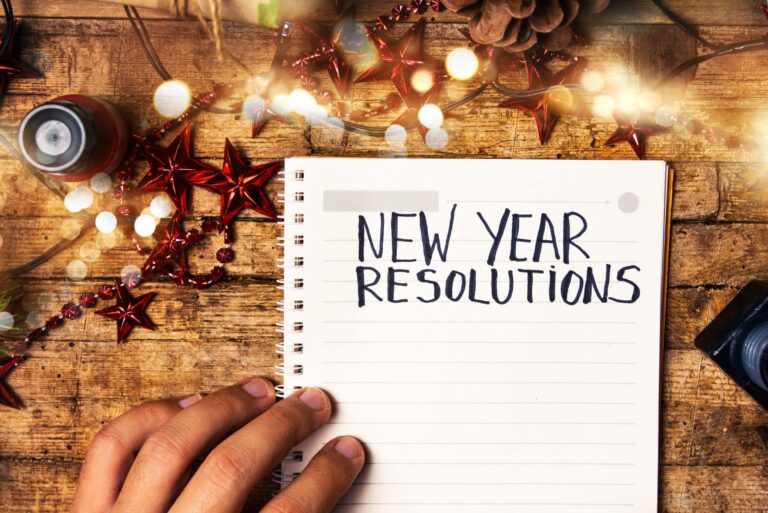 A hand is holding an open spiral notebook on a wooden table, with the words “NEW YEAR RESOLUTIONS” written in bold letters on the page. The table is decorated with festive items such as red star ornaments, pinecones, and string lights, creating a holiday-themed setting.