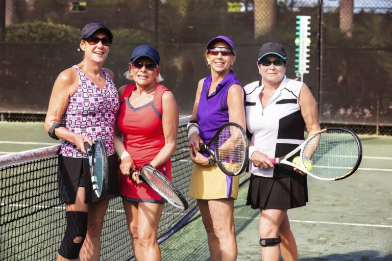 Four people standing on a tennis court near the net, each holding a tennis racket. They are wearing athletic outfits in various colors, including patterned tops, skirts, and visors. The background shows a dark fence and greenery.