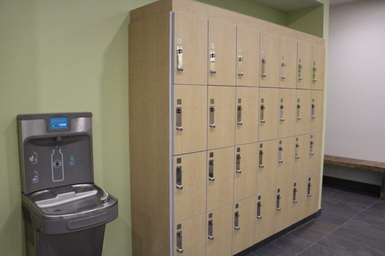Locker room area featuring a light wood locker unit with multiple compartments, each equipped with a silver handle and lock. To the left, there is a modern water bottle filling station and drinking fountain with a digital display. The walls are painted light green, and the floor is covered with dark gray tiles. A wooden bench is visible along the right wall, providing seating.