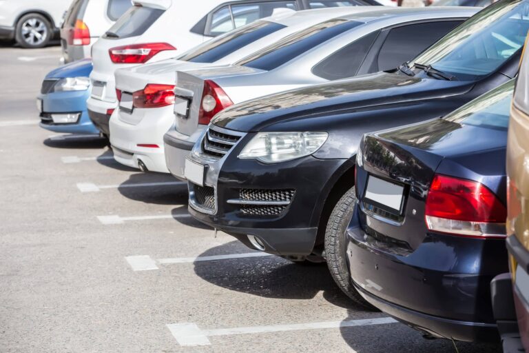 Row of parked cars in an outdoor parking lot, showing the rear ends of multiple vehicles in different colors, including black, silver, and white, lined up in marked parking spaces on asphalt.
