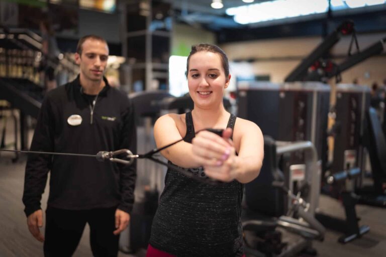 Person performing a cable exercise in a gym, holding the cable handle with both hands extended forward, while another individual stands nearby observing. The background includes various strength training machines and gym equipment.