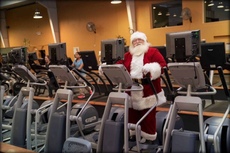 Person dressed in a full Santa Claus costume standing on an elliptical machine in a gym. The gym has multiple cardio machines arranged in rows, including treadmills and ellipticals, with other people exercising in the background. The setting features warm-colored walls, large mirrors, and overhead lighting, creating a festive and humorous scene of holiday spirit in a fitness environment.