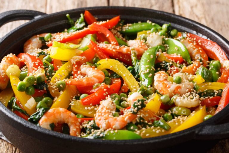 A black skillet filled with a colorful stir-fry of shrimp, sliced red and yellow bell peppers, snap peas, and green peas. The dish is topped with a generous sprinkle of sesame seeds, and the vegetables appear fresh and vibrant. The skillet is placed on a wooden surface, creating a rustic presentation that highlights the bright colors and textures of the ingredients