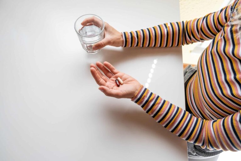 Person wearing a striped long-sleeve shirt holding a glass of water in one hand and two pills in the other, positioned over a white surface.