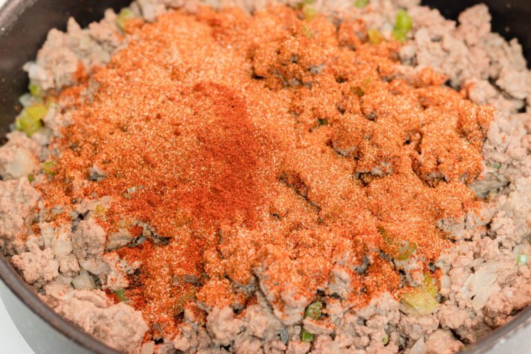 Close-up of a skillet containing cooked ground meat mixed with diced onions and green peppers, topped with a generous layer of reddish-brown seasoning powder.