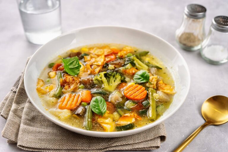 A white bowl filled with vegetable soup is placed on a beige cloth napkin on a light-colored surface. The soup contains a variety of colorful vegetables, including sliced carrots, broccoli florets, green beans, mushrooms, and leafy greens, all in a clear broth. Fresh basil leaves are used as garnish on top. Nearby, there is a gold-colored spoon, a glass of water, and two small shakers for salt and pepper, adding to the cozy and appetizing presentation.