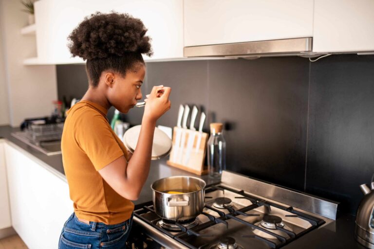 Person standing at a stove tasting food from a pot with a spoon in a modern kitchen.