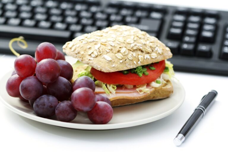Plate with a sandwich made of oat-topped bread, lettuce, tomato, and deli meat, served with a bunch of red grapes, placed near a pen and computer keyboard.